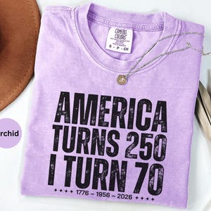 May include: Orchid-colored t-shirt with the text "AMERICA TURNS 250 I TURN 70" in black. The shirt has a crew neck and short sleeves. A silver necklace with a pendant is draped across the shirt.