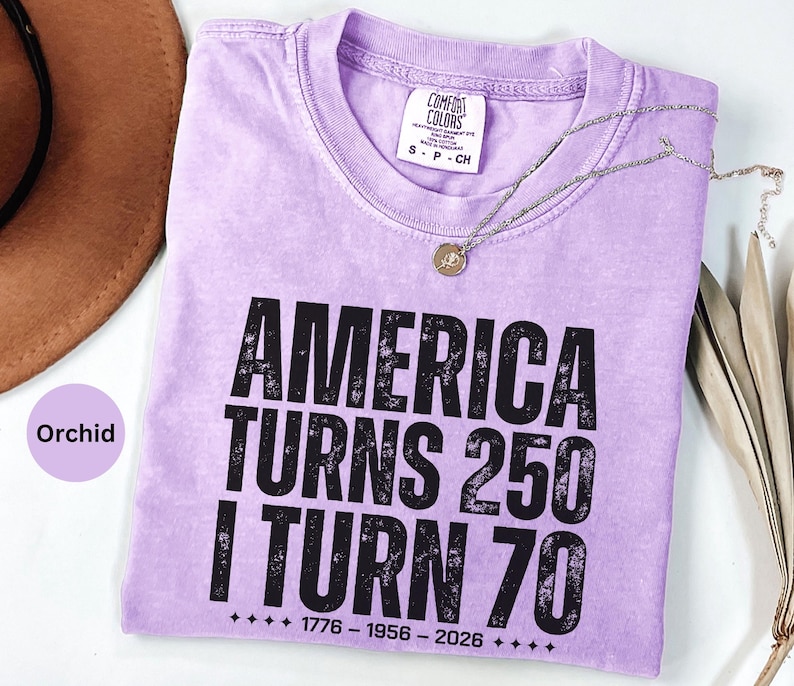 May include: Orchid-colored t-shirt with the text "AMERICA TURNS 250 I TURN 70" in black. The shirt has a crew neck and short sleeves. A silver necklace with a pendant is draped across the shirt.