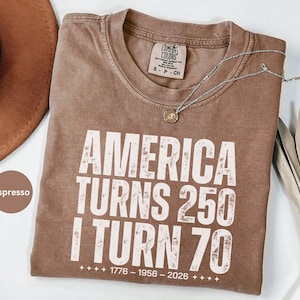 May include: Espresso-colored t-shirt with the text "AMERICA TURNS 250 I TURN 70" in white. The shirt has a crew neck and a Comfort Colors tag. A silver necklace with a pendant is draped across the shirt.