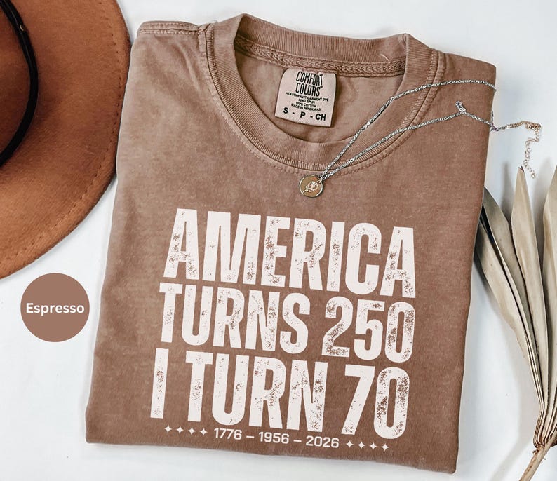 May include: Espresso-colored t-shirt with the text "AMERICA TURNS 250 I TURN 70" in white. The shirt has a crew neck and a Comfort Colors tag. A silver necklace with a pendant is draped across the shirt.