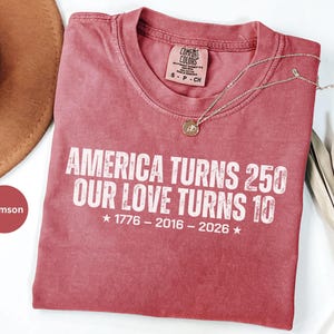May include: A crimson short-sleeve t-shirt with a crew neckline. The shirt features white distressed text that reads: AMERICA TURNS 250 OUR LOVE TURNS 10, followed by the years 1776 - 2016 - 2026 with a star on each side of the date sequence.