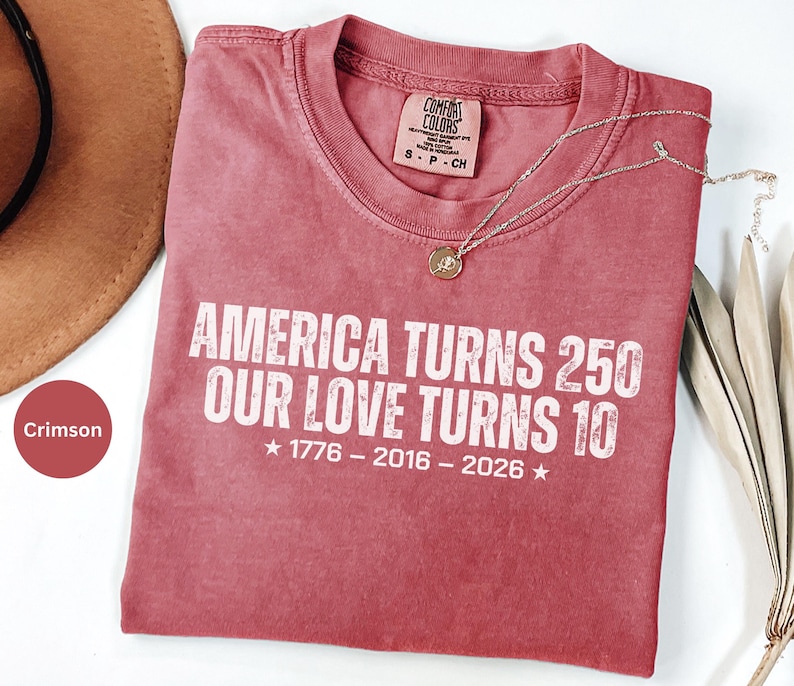 May include: A crimson short-sleeve t-shirt with a crew neckline. The shirt features white distressed text that reads: AMERICA TURNS 250 OUR LOVE TURNS 10, followed by the years 1776 - 2016 - 2026 with a star on each side of the date sequence.