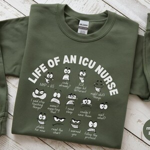 May include: Military green sweatshirt with the text "LIFE OF AN ICU NURSE" and various cartoon faces expressing different emotions. The sweatshirt also has phrases like "code already?" and "wait what?"