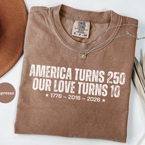 May include: An espresso-colored short-sleeve t-shirt with white distressed text that reads: AMERICA TURNS 250 OUR LOVE TURNS 10, followed by the years 1776 - 2016 - 2026 with stars on each side. The shirt features a Comfort Colors brand neck label.