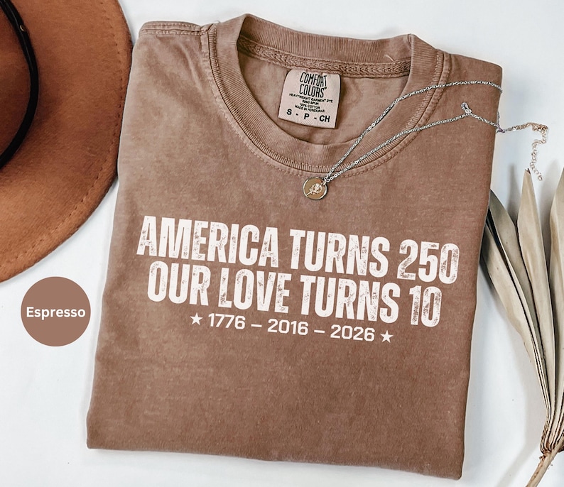 May include: An espresso-colored short-sleeve t-shirt with white distressed text that reads: AMERICA TURNS 250 OUR LOVE TURNS 10, followed by the years 1776 - 2016 - 2026 with stars on each side. The shirt features a Comfort Colors brand neck label.