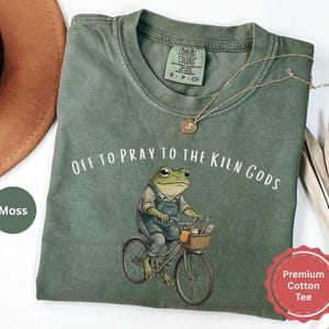 May include: Moss green premium cotton tee with the text "OFF TO PRAY TO THE KILN GODS" and a frog riding a bicycle. The frog is wearing overalls and carrying tools in a basket. A gold necklace is draped on the shirt.