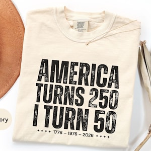 May include: Ivory-colored t-shirt with the text "AMERICA TURNS 250 I TURN 50" in distressed black lettering. The shirt also includes the years 1776-1976-2026. A tan hat and dried palm leaves are in the background.
