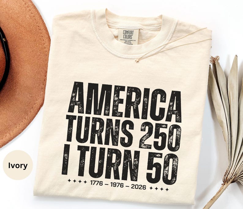 May include: Ivory-colored t-shirt with the text "AMERICA TURNS 250 I TURN 50" in distressed black lettering. The shirt also includes the years 1776-1976-2026. A tan hat and dried palm leaves are in the background.