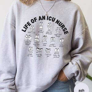 May include: Ash-colored sweatshirt with the text "LIFE OF AN ICU NURSE" in black. The design features cartoon faces with various expressions and phrases related to nursing, such as "MAP > 65" and "I need sleep."