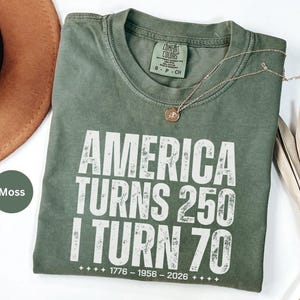 May include: A moss green t-shirt with the text "AMERICA TURNS 250 I TURN 70" in white. The shirt has a crew neck and a small tag. A gold necklace with a pendant is on the shirt.