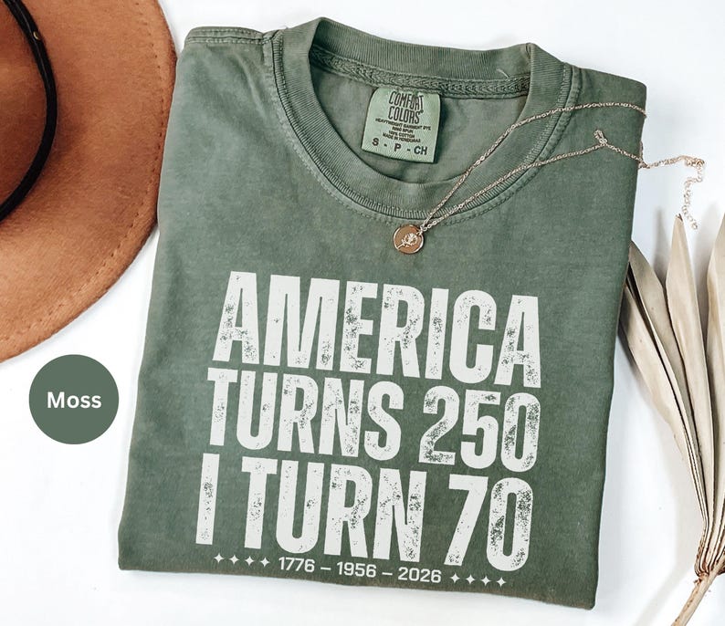 May include: A moss green t-shirt with the text "AMERICA TURNS 250 I TURN 70" in white. The shirt has a crew neck and a small tag. A gold necklace with a pendant is on the shirt.