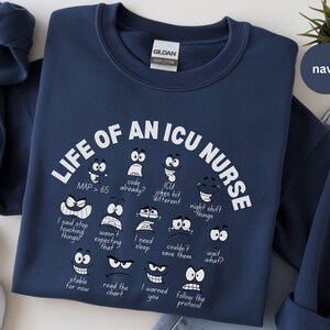 May include: Navy blue sweatshirt with the text "LIFE OF AN ICU NURSE" in white. The design includes various cartoon faces with phrases like "code already?" and "wait what?" A navy circle with the word "navy" is in the upper right.