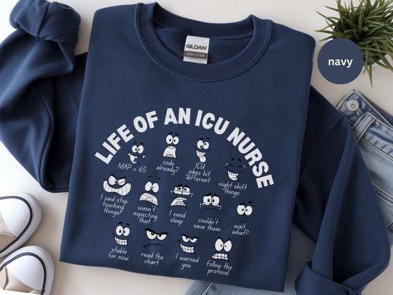 May include: Navy blue sweatshirt with the text "LIFE OF AN ICU NURSE" in white. The design includes various cartoon faces with phrases like "code already?" and "wait what?" A navy circle with the word "navy" is in the upper right.
