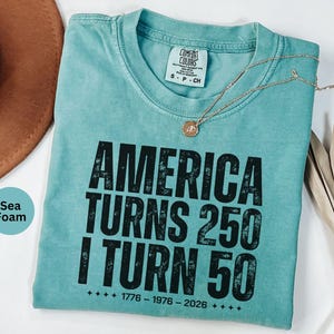 May include: Seafoam green t-shirt with the text "AMERICA TURNS 250 I TURN 50" in black. The shirt has a crew neck and a small tag at the neckline. A gold necklace with a pendant is draped across the shirt.