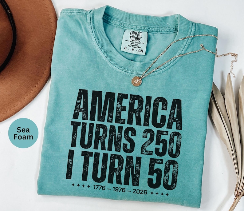May include: Seafoam green t-shirt with the text "AMERICA TURNS 250 I TURN 50" in black. The shirt has a crew neck and a small tag at the neckline. A gold necklace with a pendant is draped across the shirt.