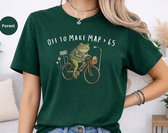 May include: A forest green short-sleeve t-shirt featuring a graphic of a frog riding a bicycle. The frog wears a medical scrub top and carries a basket. The text above the illustration reads "OFF TO MAKE MAP > 65" in white capital letters.