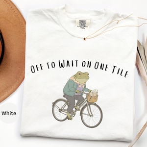 Op de afbeelding: Een wit t-shirt met een afbeelding van een groene kikker met een sjaal, jas en broek op een fiets met een mandje. De tekst "OFF TO WAIT ON ONE TILE" staat in het zwart boven de illustratie van de kikker. Het shirt heeft een ronde hals.