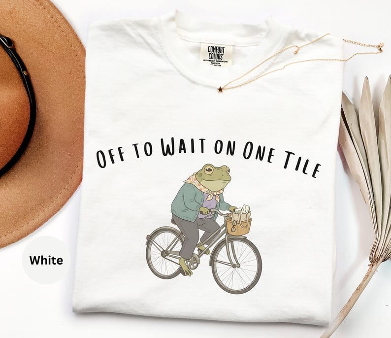 Op de afbeelding: Een wit t-shirt met een afbeelding van een groene kikker met een sjaal, jas en broek op een fiets met een mandje. De tekst "OFF TO WAIT ON ONE TILE" staat in het zwart boven de illustratie van de kikker. Het shirt heeft een ronde hals.