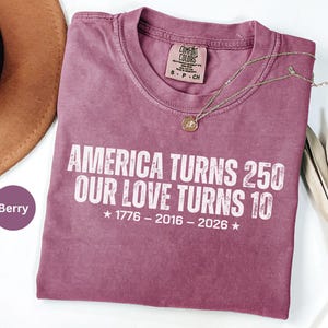 May include: A berry-colored short-sleeve t-shirt with white text that reads AMERICA TURNS 250 OUR LOVE TURNS 10, followed by the years 1776 - 2016 - 2026 with stars on each side. The shirt features a Comfort Colors brand label in the neck area.