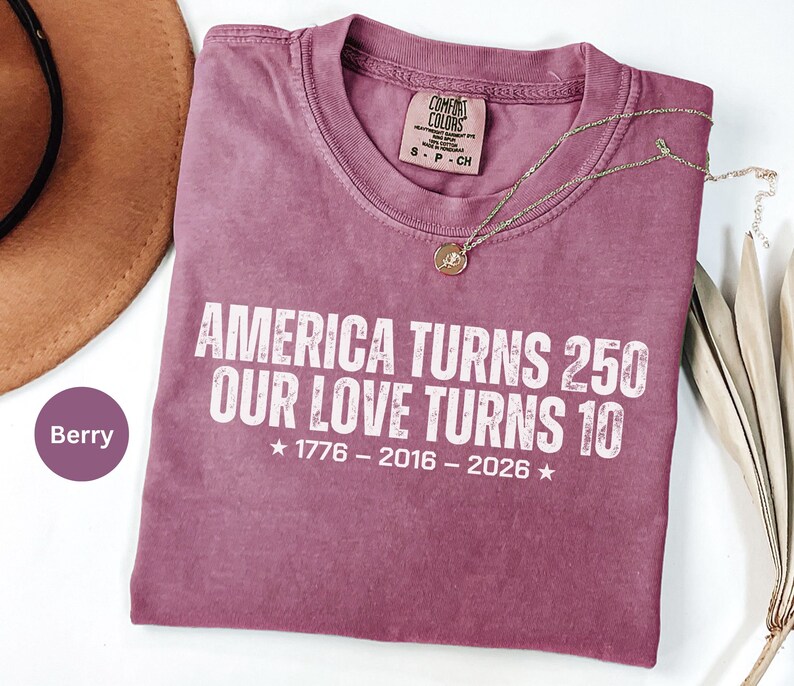 May include: A berry-colored short-sleeve t-shirt with white text that reads AMERICA TURNS 250 OUR LOVE TURNS 10, followed by the years 1776 - 2016 - 2026 with stars on each side. The shirt features a Comfort Colors brand label in the neck area.