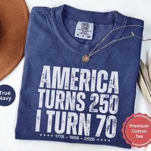 May include: A navy blue t-shirt with the text "AMERICA TURNS 250 I TURN 70" in white. The shirt has a crew neck and short sleeves. A gold necklace with a pendant is draped across the shirt.