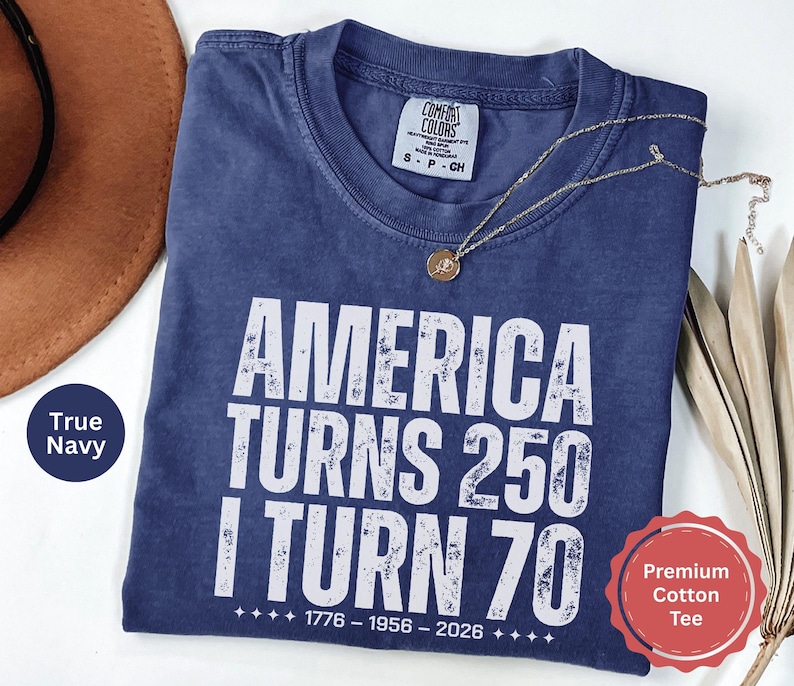 May include: A navy blue t-shirt with the text "AMERICA TURNS 250 I TURN 70" in white. The shirt has a crew neck and short sleeves. A gold necklace with a pendant is draped across the shirt.