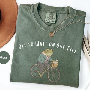 Op de afbeelding: Een mosgroen t-shirt met ronde hals en een afbeelding van een kikker in een vest en sjaal op een fiets met een mandje. De tekst "OFF TO WAIT ON ONE TILE" staat in het wit boven de kikker. Het shirt heeft een Comfort Colors label.