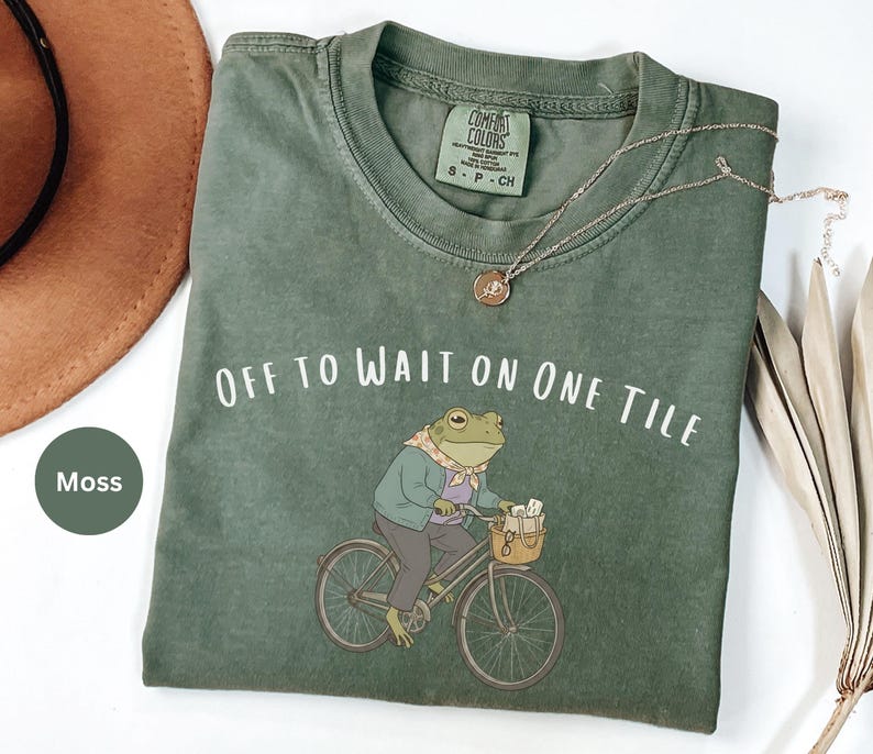Op de afbeelding: Een mosgroen t-shirt met ronde hals en een afbeelding van een kikker in een vest en sjaal op een fiets met een mandje. De tekst "OFF TO WAIT ON ONE TILE" staat in het wit boven de kikker. Het shirt heeft een Comfort Colors label.