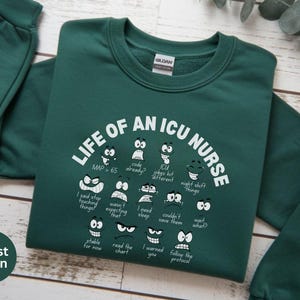 May include: Forest green sweatshirt with the text "LIFE OF AN ICU NURSE" in white, along with various cartoon faces expressing different emotions. The sweatshirt is made of a soft material.