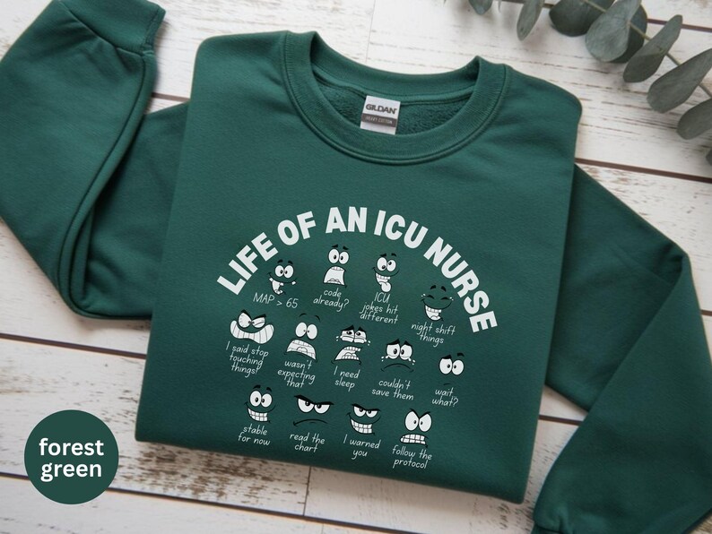 May include: Forest green sweatshirt with the text "LIFE OF AN ICU NURSE" in white, along with various cartoon faces expressing different emotions. The sweatshirt is made of a soft material.