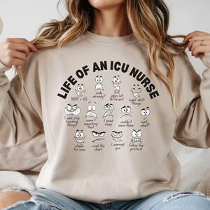 May include: Sand-colored sweatshirt with the text "LIFE OF AN ICU NURSE" in black, featuring cartoon faces expressing various emotions. The sweatshirt also includes phrases like "MAP > 65" and "I said stop touching things!"
