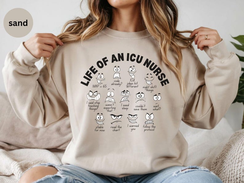 May include: Sand-colored sweatshirt with the text "LIFE OF AN ICU NURSE" in black, featuring cartoon faces expressing various emotions. The sweatshirt also includes phrases like "MAP > 65" and "I said stop touching things!"