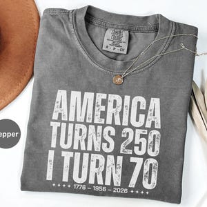May include: A gray Comfort Colors t-shirt with the text "AMERICA TURNS 250 I TURN 70" in white block letters. The shirt also has the dates 1776-1956-2026 printed below the text. A gold necklace with a pendant is draped on the shirt.