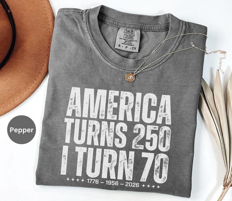 May include: A gray Comfort Colors t-shirt with the text "AMERICA TURNS 250 I TURN 70" in white block letters. The shirt also has the dates 1776-1956-2026 printed below the text. A gold necklace with a pendant is draped on the shirt.