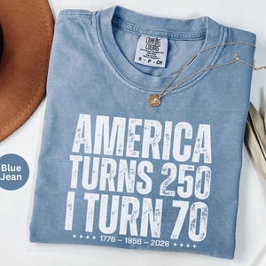 May include: Blue jean colored t-shirt with the text "AMERICA TURNS 250 I TURN 70" in white. The shirt has a crew neck and a small tag on the collar. A gold necklace and a brown hat are visible.
