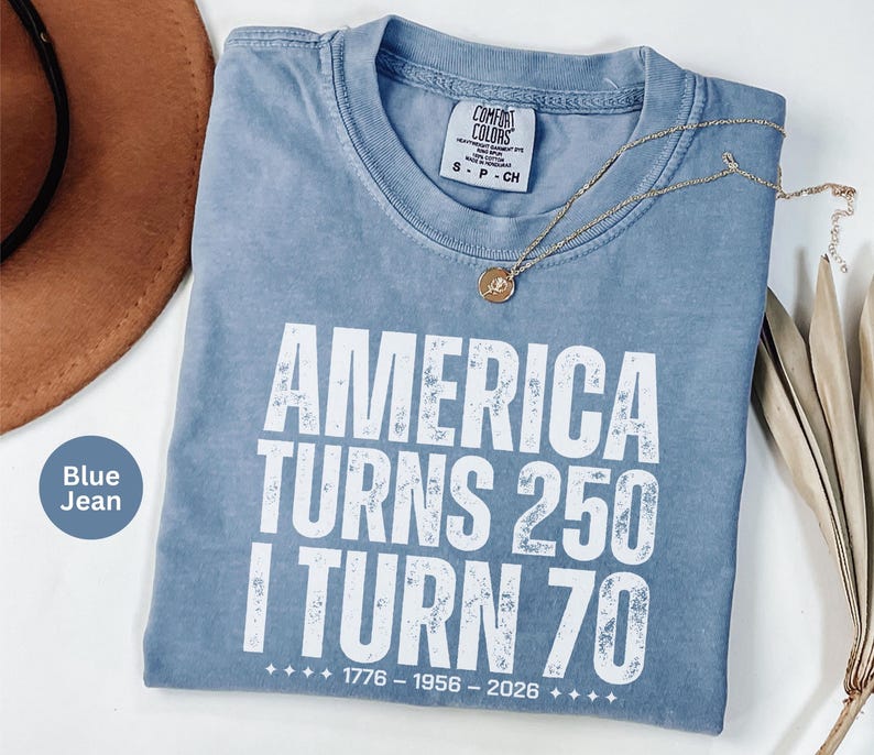 May include: Blue jean colored t-shirt with the text "AMERICA TURNS 250 I TURN 70" in white. The shirt has a crew neck and a small tag on the collar. A gold necklace and a brown hat are visible.
