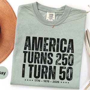 May include: A light green t-shirt with the text "AMERICA TURNS 250 I TURN 50" in black. The shirt also has the dates 1776-1976-2026 printed on it. A brown hat is visible in the background.
