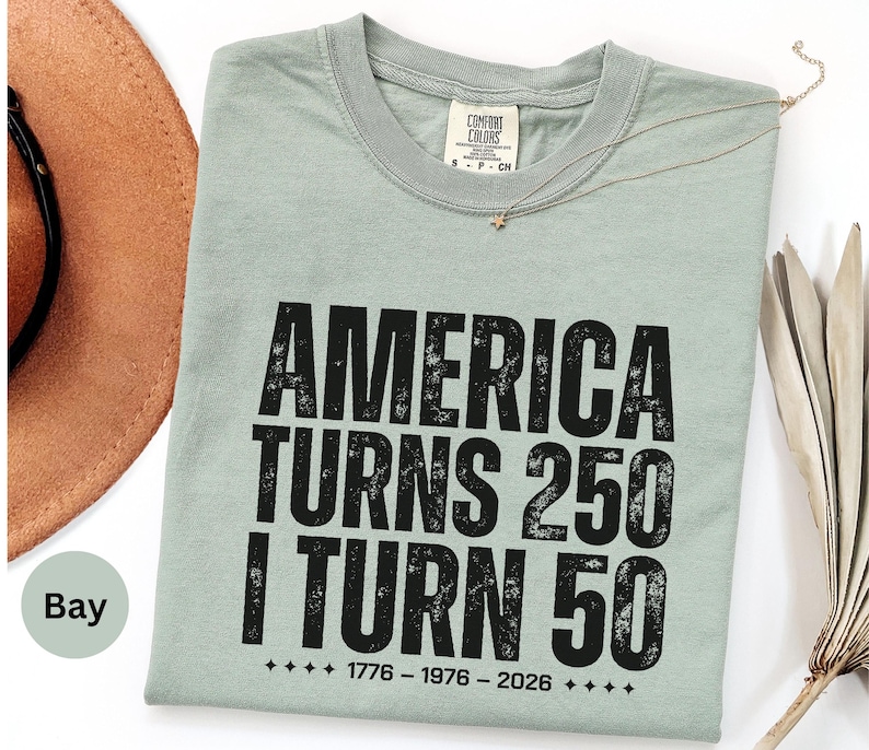 May include: A light green t-shirt with the text "AMERICA TURNS 250 I TURN 50" in black. The shirt also has the dates 1776-1976-2026 printed on it. A brown hat is visible in the background.
