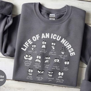 May include: Graphite heather sweatshirt with the text "LIFE OF AN ICU NURSE" and various cartoon faces expressing different emotions. The sweatshirt has long sleeves and a crew neck. The text and graphics are in white.