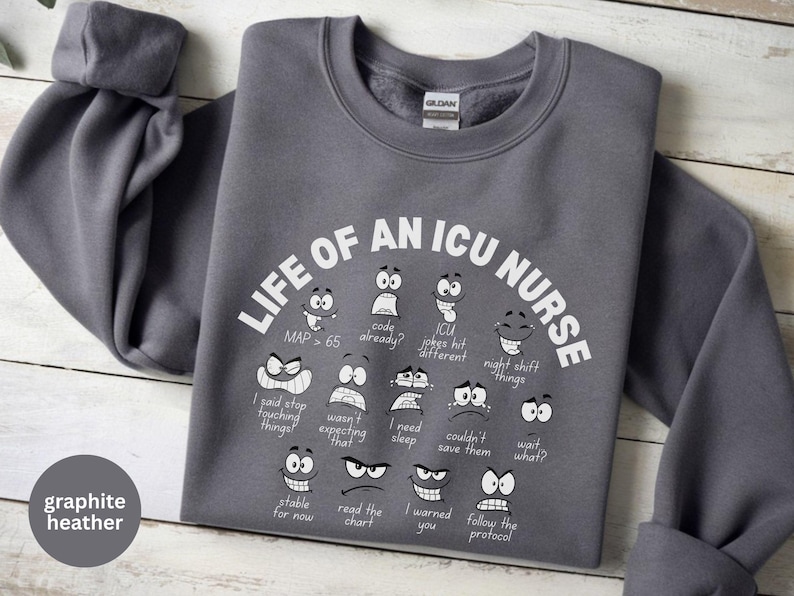 May include: Graphite heather sweatshirt with the text "LIFE OF AN ICU NURSE" and various cartoon faces expressing different emotions. The sweatshirt has long sleeves and a crew neck. The text and graphics are in white.