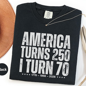 May include: Black t-shirt with white distressed text: "AMERICA TURNS 250 I TURN 70" and dates 1776-1956-2026. A gold star necklace and a brown hat are visible. The shirt is a Comfort Colors brand.
