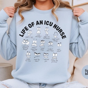 May include: Light blue sweatshirt with the text "LIFE OF AN ICU NURSE" and various cartoon faces with different expressions. The sweatshirt also has the text "MAP > 65".