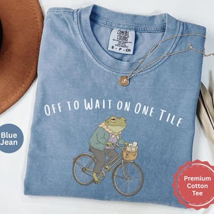 Op de afbeelding: Een blauw spijkerkleurig t-shirt met een afbeelding van een kikker met een sjaal en jas op een fiets. De tekst boven de kikker luidt "OFF TO WAIT ON ONE TILE". Het t-shirt heeft een Comfort Colors merklabel bij de hals en korte mouwen.