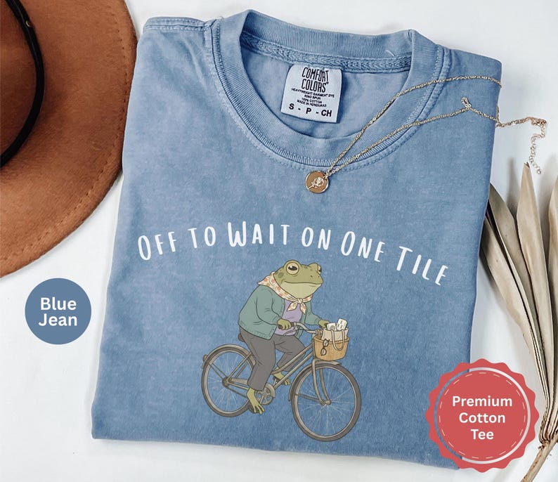 Op de afbeelding: Een blauw spijkerkleurig t-shirt met een afbeelding van een kikker met een sjaal en jas op een fiets. De tekst boven de kikker luidt "OFF TO WAIT ON ONE TILE". Het t-shirt heeft een Comfort Colors merklabel bij de hals en korte mouwen.