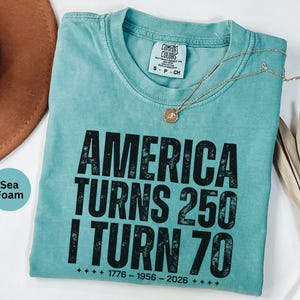 May include: Seafoam green t-shirt with the text "AMERICA TURNS 250 I TURN 70" in black. The shirt has a crew neck and a small tag at the neckline. A gold necklace with a pendant is draped across the shirt.