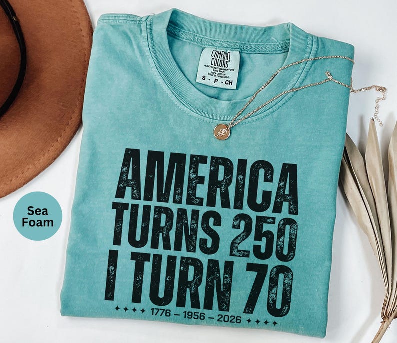 May include: Seafoam green t-shirt with the text "AMERICA TURNS 250 I TURN 70" in black. The shirt has a crew neck and a small tag at the neckline. A gold necklace with a pendant is draped across the shirt.