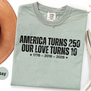 May include: A sage green short-sleeve t-shirt with black distressed text that reads: AMERICA TURNS 250 OUR LOVE TURNS 10, followed by the years 1776 - 2016 - 2026 with a star on each side. The shirt features a crew neckline and a Comfort Colors label.