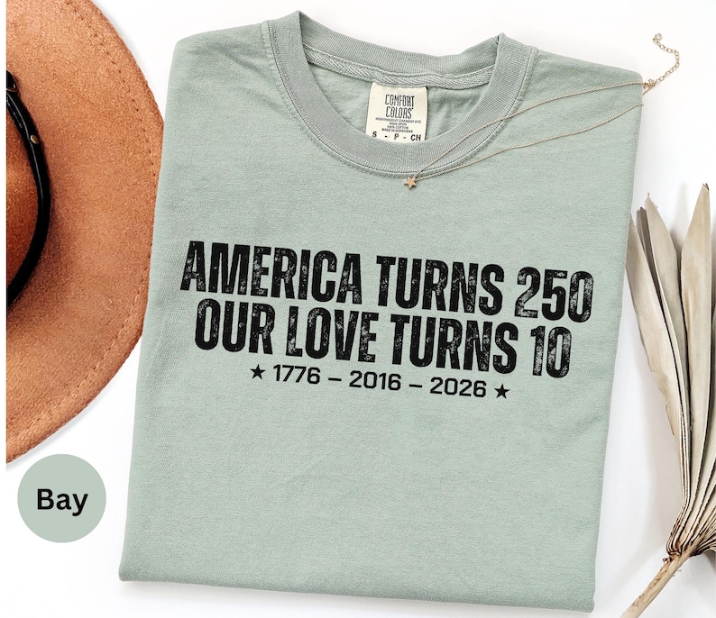 May include: A sage green short-sleeve t-shirt with black distressed text that reads: AMERICA TURNS 250 OUR LOVE TURNS 10, followed by the years 1776 - 2016 - 2026 with a star on each side. The shirt features a crew neckline and a Comfort Colors label.