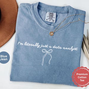 May include: A blue jean colored short-sleeve t-shirt with a crew neckline. The shirt features the white text I'm literally just a data analyst above a white line drawing of a ribbon bow. A gold-toned necklace with a small circular pendant rests on top.