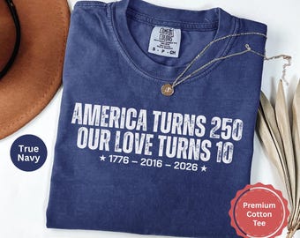 May include: A navy blue short-sleeved cotton t-shirt with white distressed text that reads AMERICA TURNS 250 OUR LOVE TURNS 10 1776 - 2016 - 2026. The shirt features a crew neckline and a Comfort Colors brand label on the inside collar area.